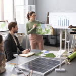 A team of professionals in a modern office discusses environmental planning and fiscal strategy. One woman presents a topographic map while another man notes data shown on a bar chart. Solar panel models and wind turbines on the table symbolize the integration of green energy with fiscal sustainability goals.