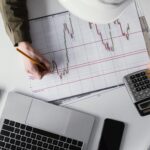 A person wearing a hard hat analyzes a financial chart with candlestick patterns while holding a calculator. The workspace includes a laptop, smartphone, notebook, and a solar panel, representing investment strategy discussions possibly related to infrastructure and the pension system.