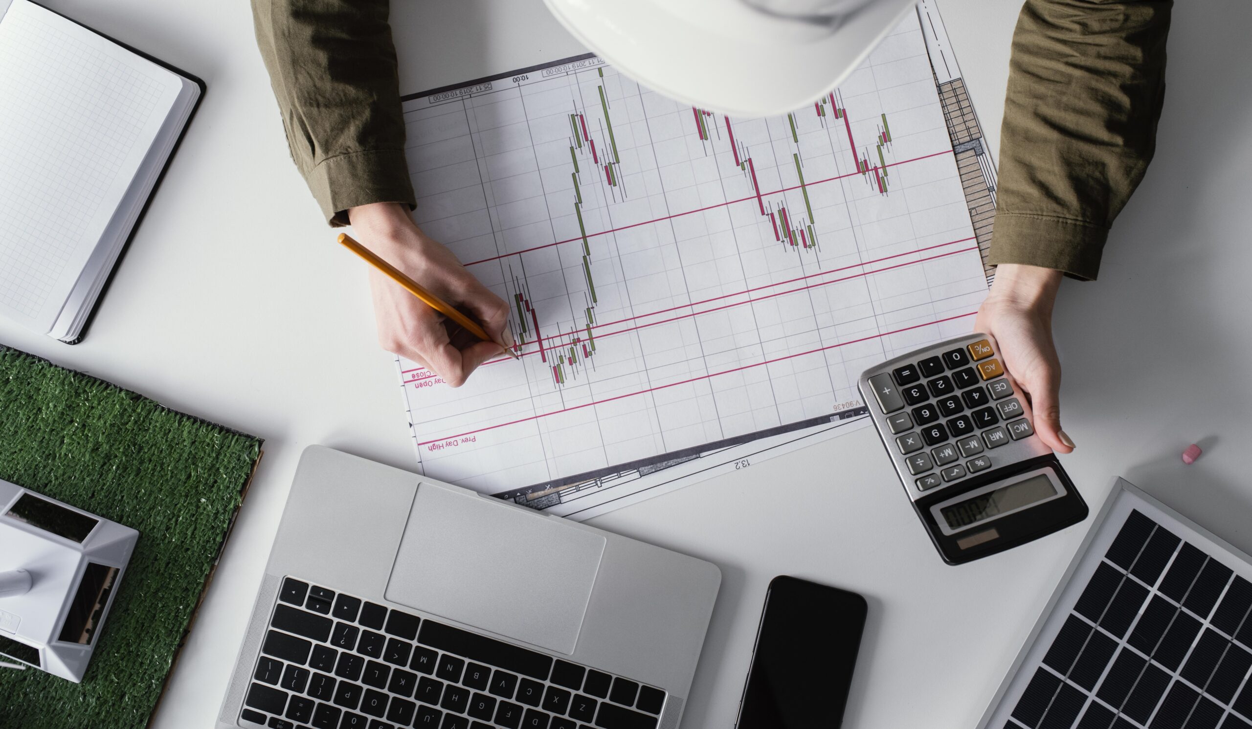 A person wearing a hard hat analyzes a financial chart with candlestick patterns while holding a calculator. The workspace includes a laptop, smartphone, notebook, and a solar panel, representing investment strategy discussions possibly related to infrastructure and the pension system.