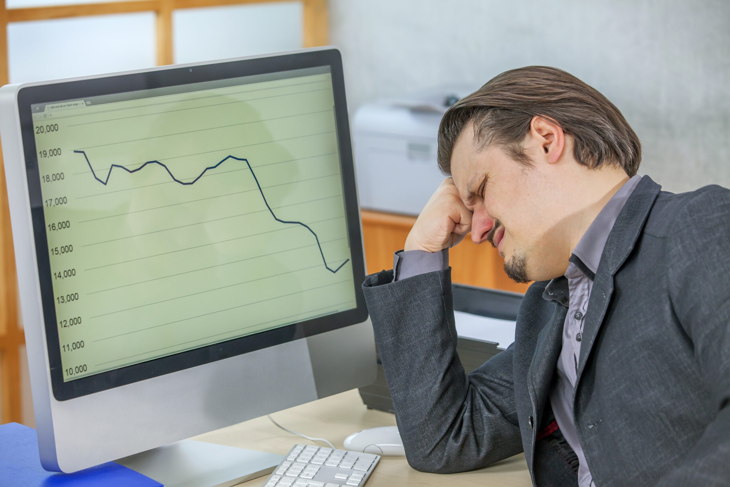 Frustrated businessman sitting at desk with head in hand while looking at a sharp decline in a stock market graph on his computer screen, symbolizing the economic impact of falling interest rates.